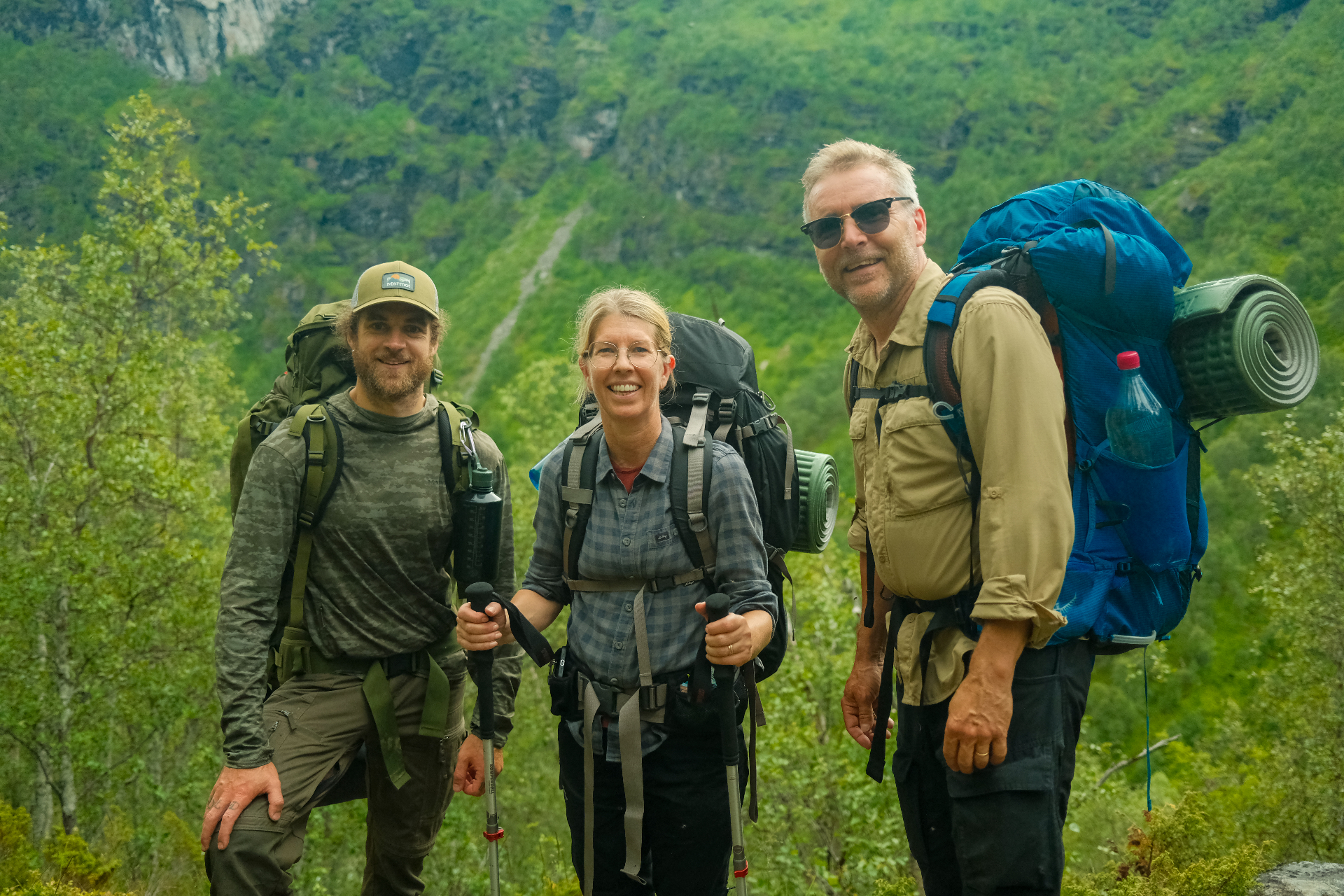 Three guests smiling with backpacks and trekking poles on a green fjord-side trail