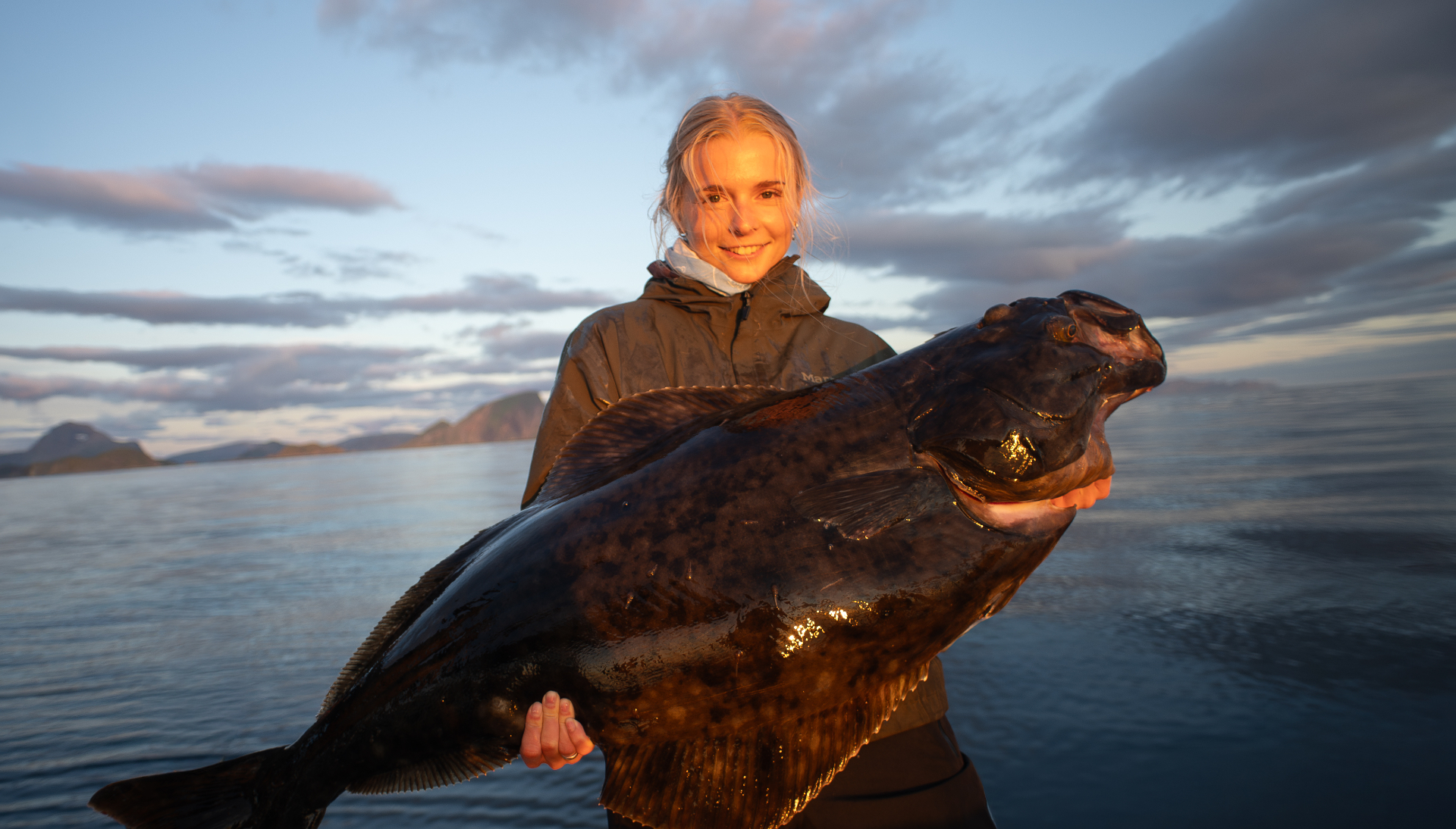 Guest holding a large halibut on the fjord under a soft Arctic-summer sky