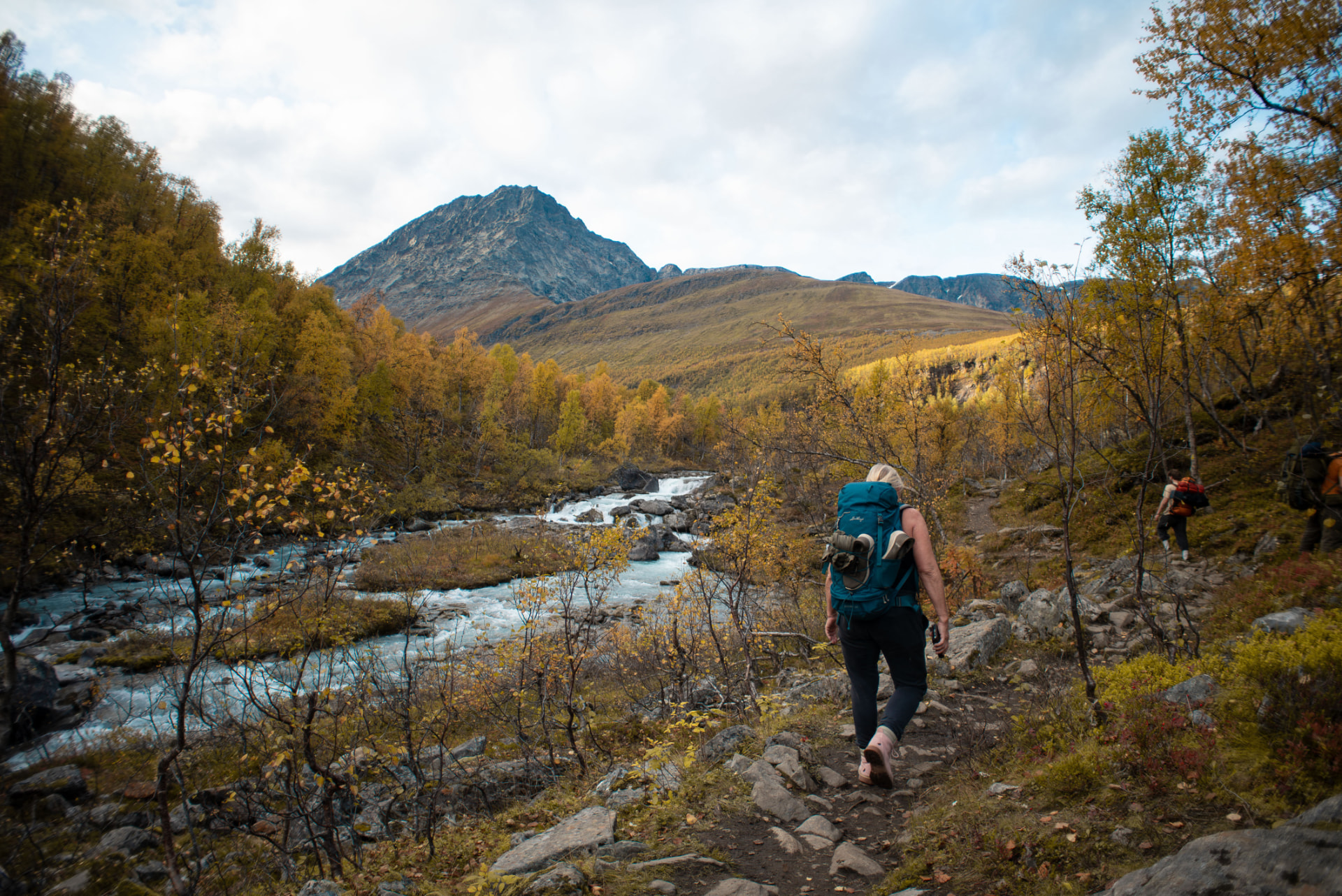 Hiker on a trail by a glacial river with a waterfall and mountain peaks beyond