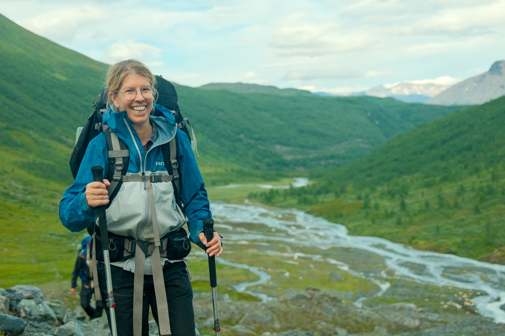 Hiker smiling with trekking poles above a braided river delta in a green Arctic valley