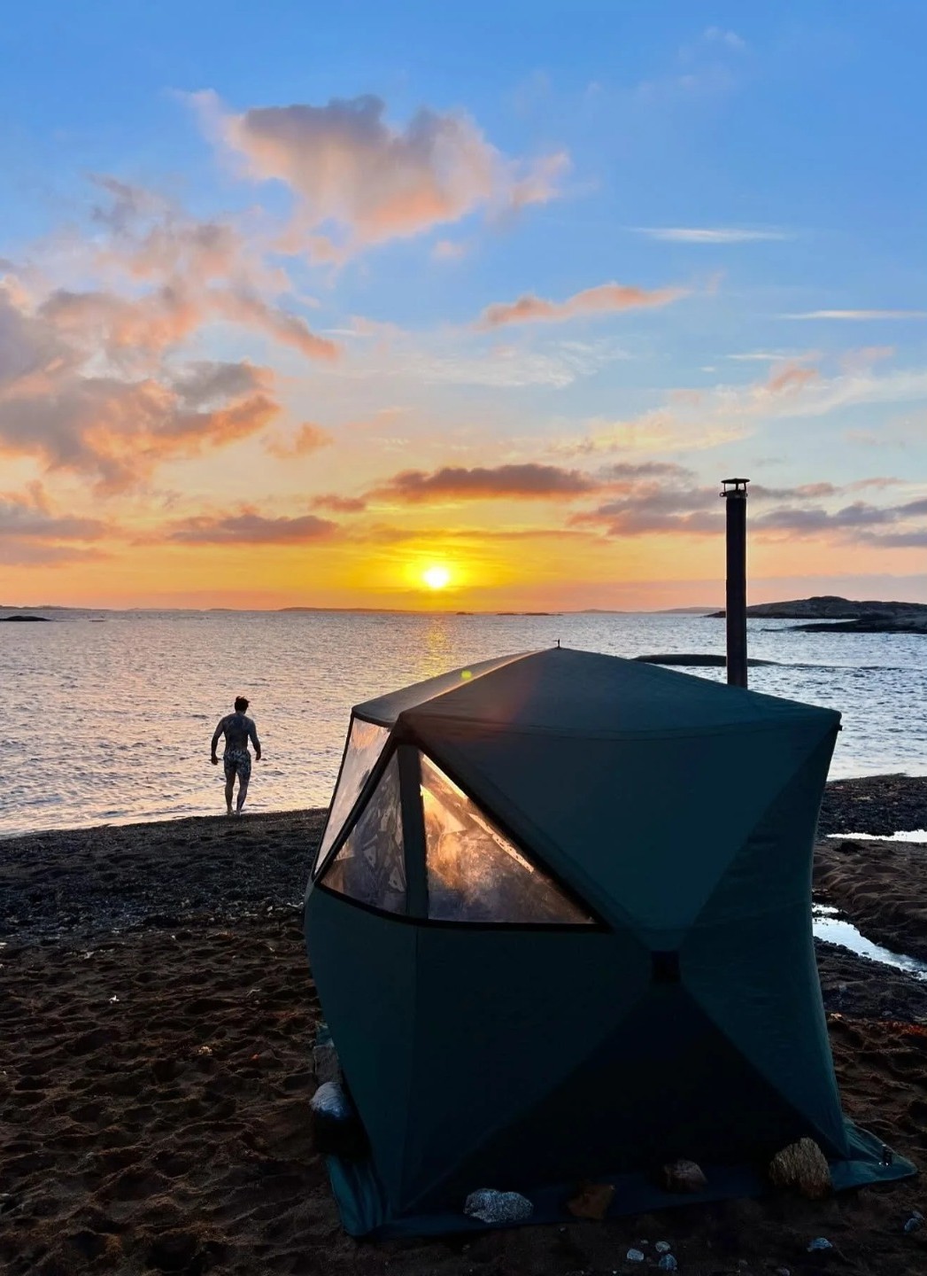 Wood-fired sauna tent on a shoreline at sunset with a swimmer walking into the fjord