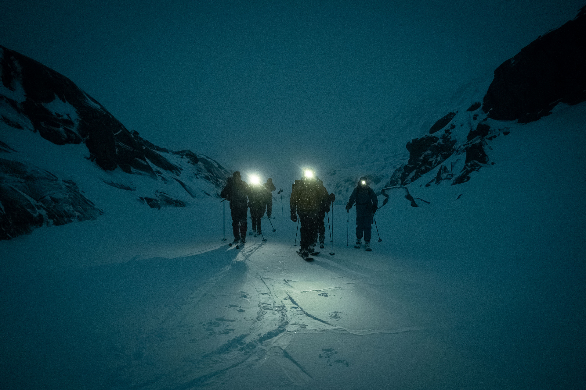 Ski tourers ascending a snowy couloir under headlamp at blue hour